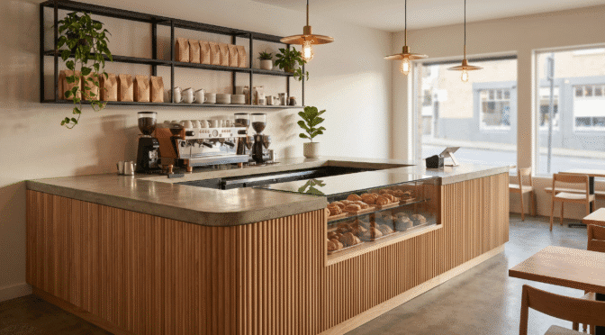 A modern coffee shop counter design featuring a durable concrete countertop, warm fluted wood paneling, and an integrated glass pastry display case.