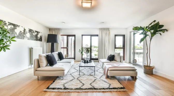 Modern Scandinavian living room with light wood floors, large windows, neutral sofas, and a patterned rug, featuring natural light and minimalist decor.
